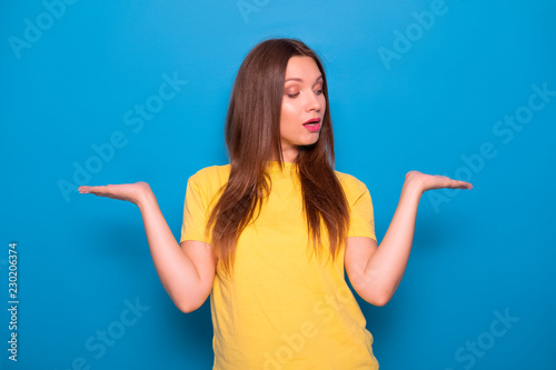 Cute brunette woman with long hair posing in yellow t-shirt on a blue background. Emotional portrait. She holds her hands in doubt what to choose
