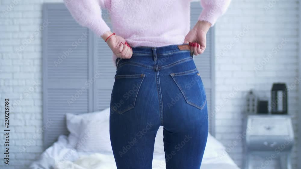 Rear view of adult woman with perfect body and buttocks fitting in tight jeans in bedroom after gaining weight. Midsection of female hands unable to close the pants due to gaining fat on hips at home.