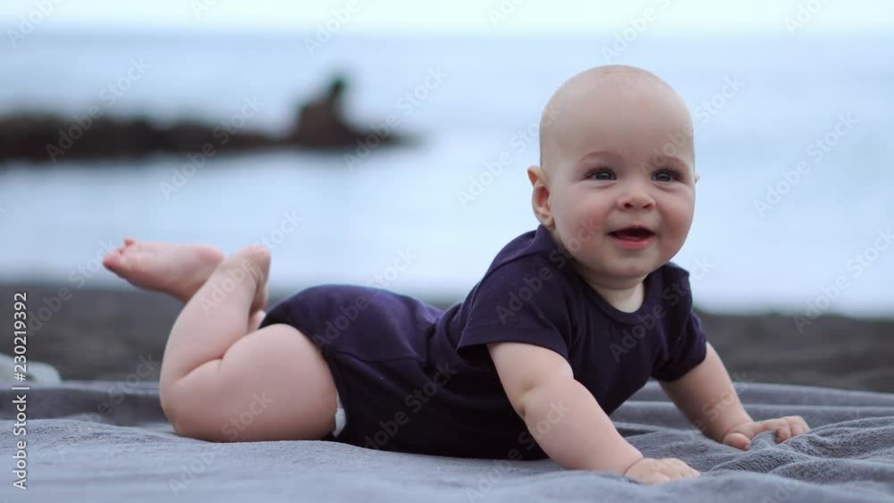 The baby lies on his stomach on the black sand near the ocean and laughs looking at the camera The baby lies on his stomach on the black sand near the ocean and laughs looking at the camera