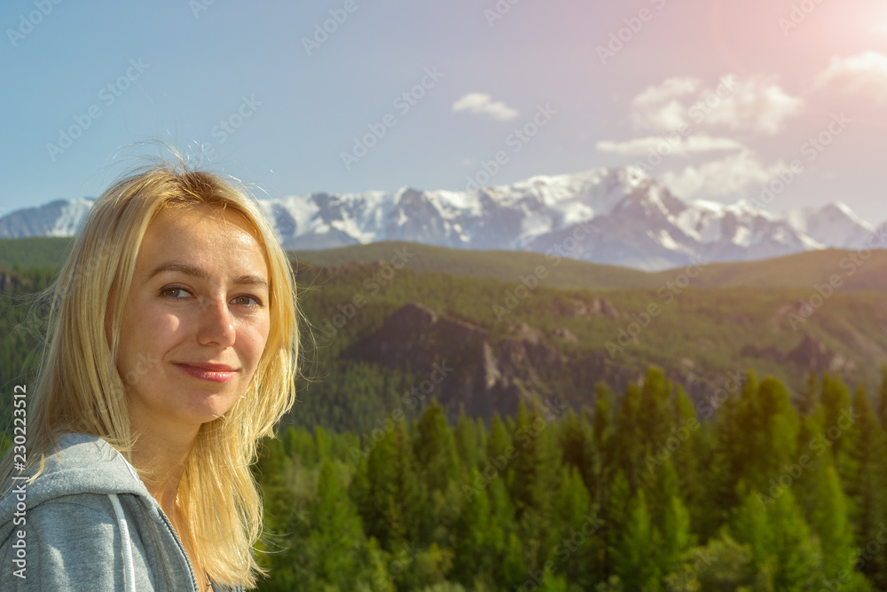 Portrait of a young beautiful blonde who smiles and looks into the camera against the backdrop of the large snow-covered Altai mountains on a clear summer day Portrait of a young beautiful blonde who smiles and looks into the camera against the backdrop of the large snow-covered Altai mountains on a clear summer day