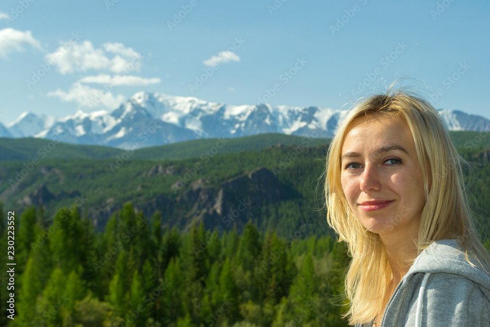 Portrait of a young beautiful blonde who smiles and looks into the camera against the backdrop of the large snow-covered Altai mountains on a clear summer day
