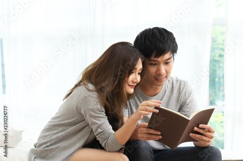 Young asian romantic couple reading book having great time together feeling satisfaction and positive in white bedroom. Close up view of happy lifestyle married couple in leisure family time concept.