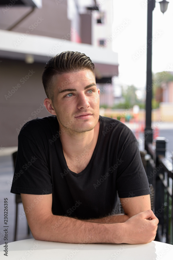 Young handsome man thinking while leaning on table sitting outsi Young handsome man thinking while leaning on table sitting outsi