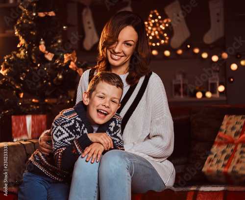 Mom hugging her cute little boy while sitting on sofa in decorated room during Christmas time. © Fxquadro
