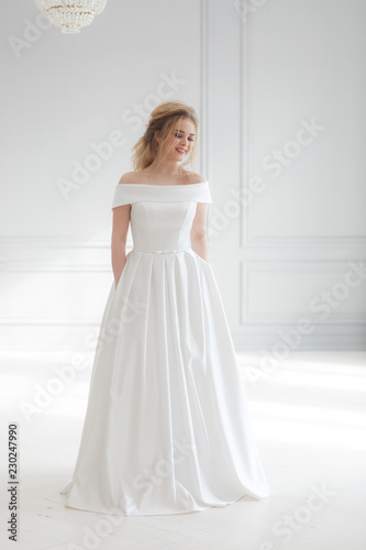 Young beautiful bride wearing white wedding dress in light classic studio interior. Vertical full-lenght portrait