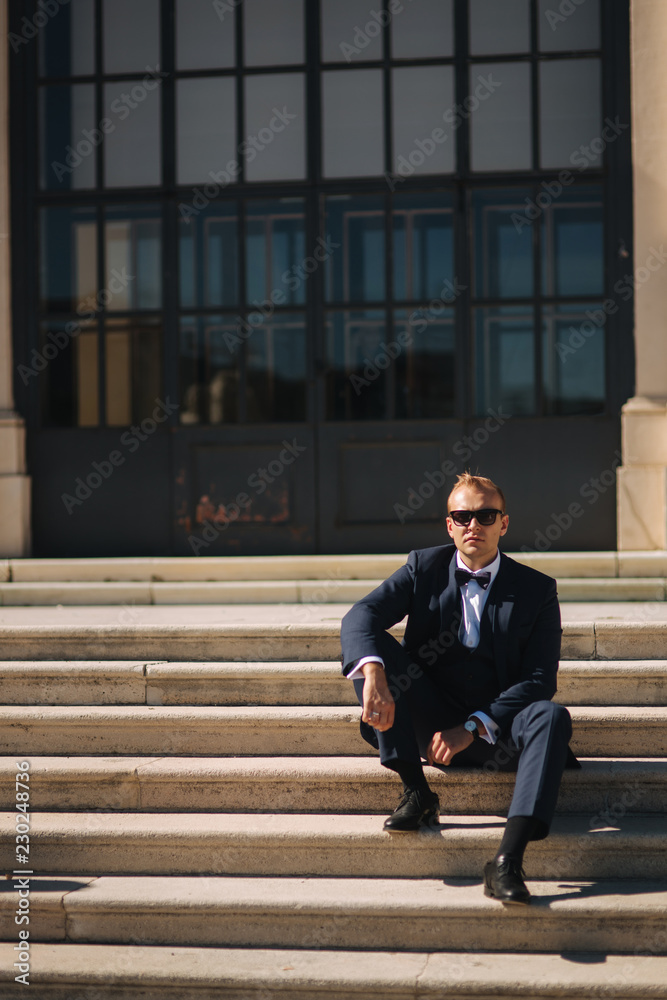 Fashion man in stylish blue suit and tie bow. He also wears sunglasses and and luxury watch