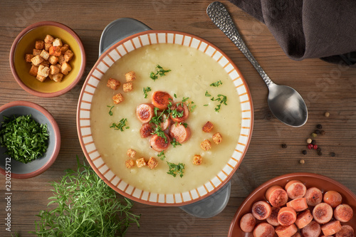 Flat lay of homemade cream of potato soup with croutons, fried vienna sausages and green coriander on wood photo