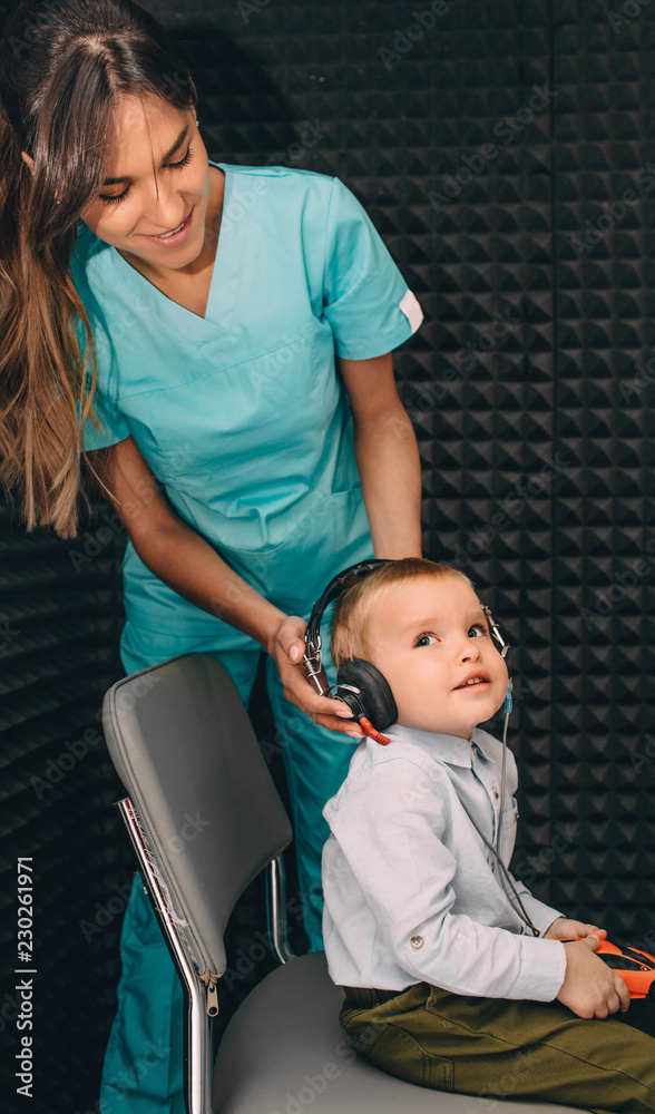 audiologist prepares little boy to hearing exam using headphones in special audio room. audiologist prepares little boy to hearing exam using headphones in special audio room.