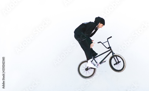 Young man in casual clothing makes tricks on bmx against the background of a white wall. BMX rider makes the Barzpin trick against its white background