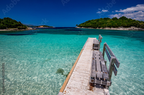 Spring daytime beautiful Ionian Sea with clear turquoise water, wooden pier and fine sand coast view from Ksamil beach, Albania. Deep blue sky with white clouds.