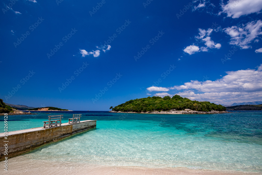 Spring daytime beautiful Ionian Sea with clear turquoise water, wooden pier and fine sand coast view from Ksamil beach, Albania. Deep blue sky with white clouds. Spring daytime beautiful Ionian Sea with clear turquoise water, wooden pier and fine sand coast view from Ksamil beach, Albania. Deep blue sky with white clouds.
