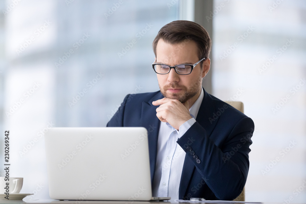 Caucasian man sitting at the table near laptop in the office and looking at the screen. Serious and pensive boss or employee working at his workplace and feeling interest or analyzing results. Caucasian man sitting at the table near laptop in the office and looking at the screen. Serious and pensive boss or employee working at his workplace and feeling interest or analyzing results.