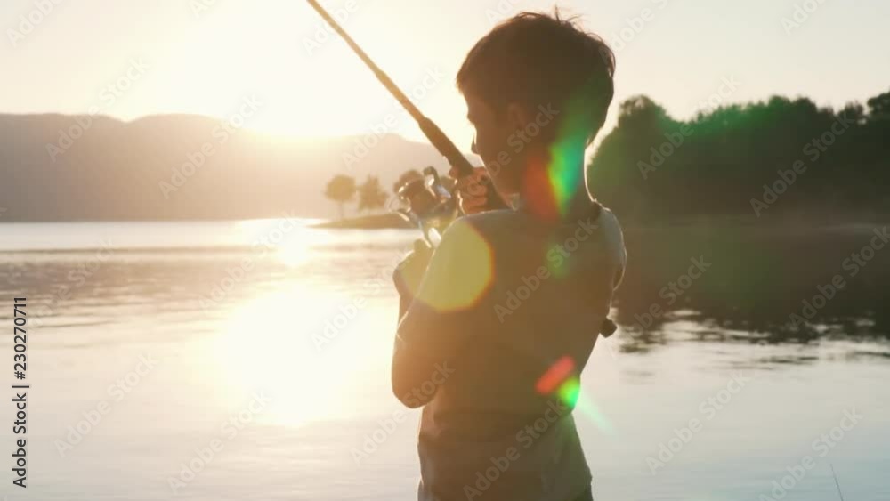 Slow motion slide fragment of rotation of the handle with a reel of fishing rod against the background of an orange sunset. A boy fishing large lake tightens the line with a reel of fish. Lens flare