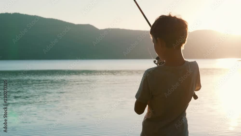 Slow motion slide fragment of rotation of the handle with a reel of fishing rod against the background of an orange sunset. A boy fishing large lake tightens the line with a reel of fish. Lens flare