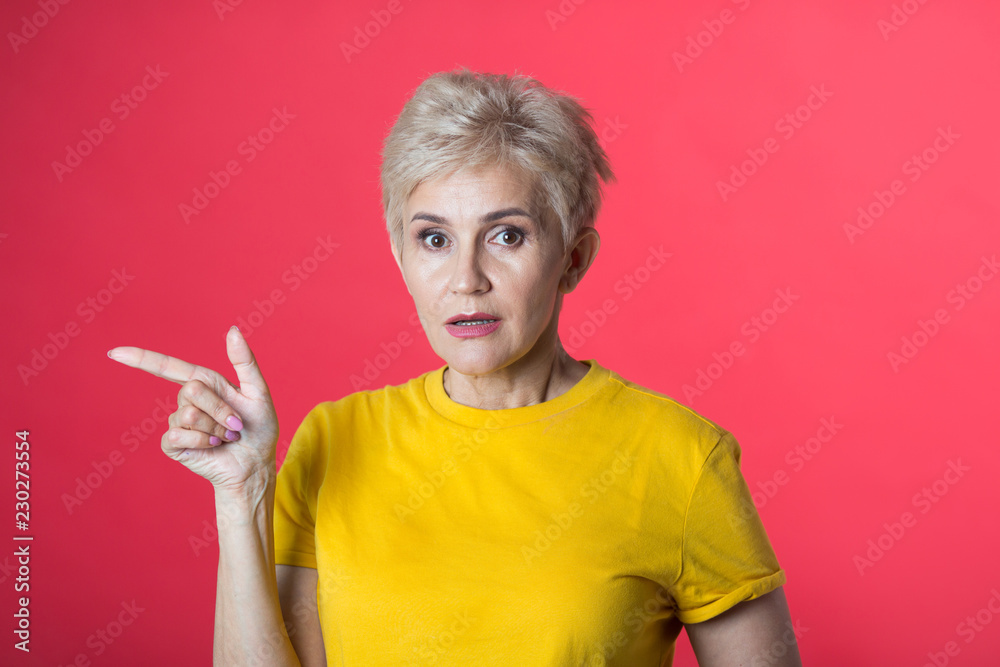stylish woman at the age with a short haircut in a yellow T-shirt on a red background with a gesture of hands stylish woman at the age with a short haircut in a yellow T-shirt on a red background with a gesture of hands