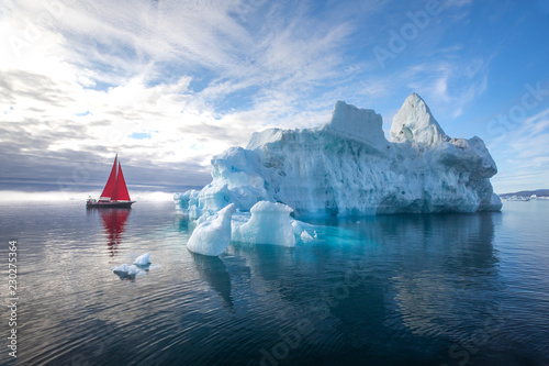 Beautiful red sailboat in the arctic next to a massive iceberg showing the scale. Ilulissat, Disko Bay, Greenland.