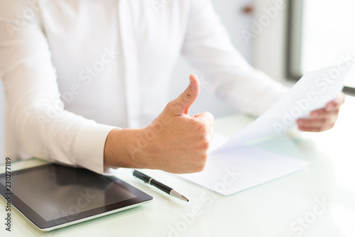 Closeup of man holding document and showing thumb up. Entrepreneur sitting at desk and working. Success and contract concept. Cropped view.