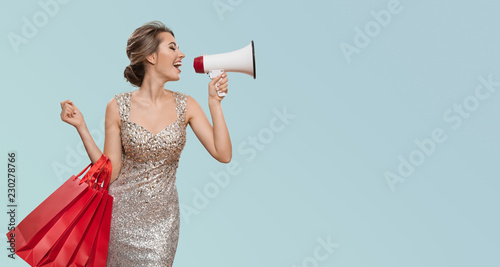 Portrait of happy charming woman holding red shopping bags. Shout into megaphone on copyspace. Blue background.