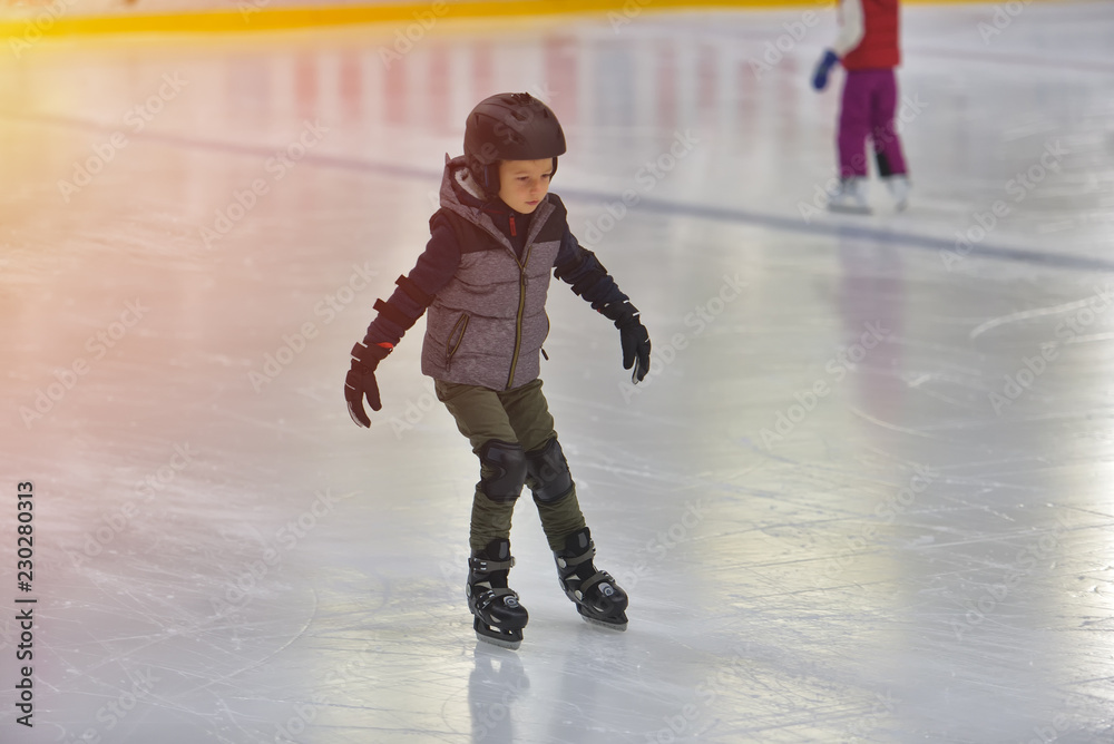 Obraz premium Adorable little boy in winter clothes with protections skating on ice rink