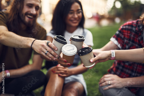 Clanging glasses. Happy friendly young people relaxing outdoors on the grass and putting their cups of hot tasty coffee together