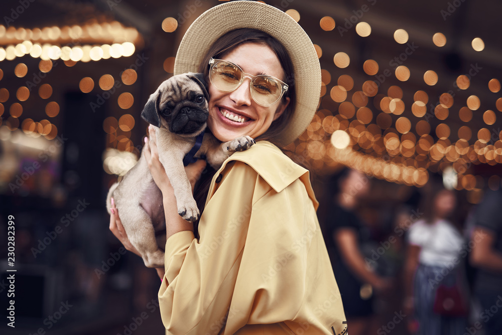 Lady with puppy. Cute pretty young lady wearing stylish hat and smiling while hugging adorable little puppy - obrazy, fototapety, plakaty 