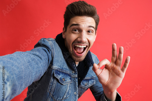 Excited young man in jeans denim jacket take selfie by camera isolated over red background showing okay gesture.