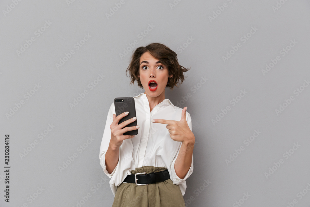 Photo of shocked woman 20s holding and using mobile phone, isolated over gray background Photo of shocked woman 20s holding and using mobile phone, isolated over gray background