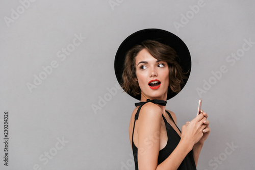 Photo of european woman 20s wearing black dress and hat smiling at camera while holding smartphone, isolated over gray background