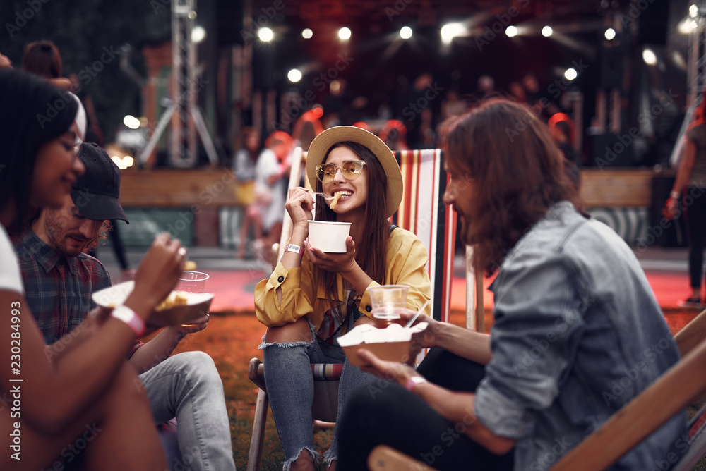 Portrait of cheerful young lady in hat sitting on folding chair and eating french fries while handsome bearded guys holding beer. Stage and crowd on blurred background