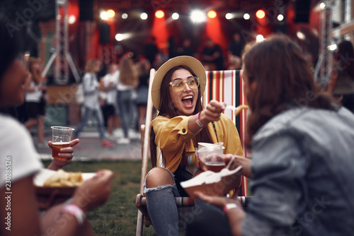 Portrait of smiling young lady in hat sitting on folding chair and letting boyfriend taste french fry. Young people resting during outdoor concert