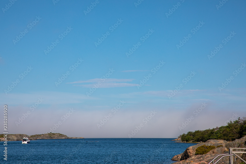 Looking towards a Baltic inlet in Kökar, a municipality of the Åland Islands, Finland, under a soft cloudy blue sky with a dark haze rolling in, during mid-morning near midsummer. Looking towards a Baltic inlet in Kökar, a municipality of the Åland Islands, Finland, under a soft cloudy blue sky with a dark haze rolling in, during mid-morning near midsummer.