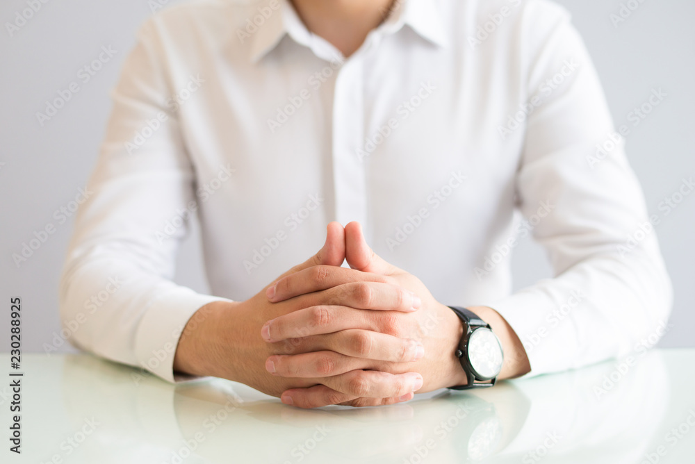 Closeup of man sitting at table with his hands clasped. Male person wearing shirt and watch. Concentration or waiting concept. Cropped front view.