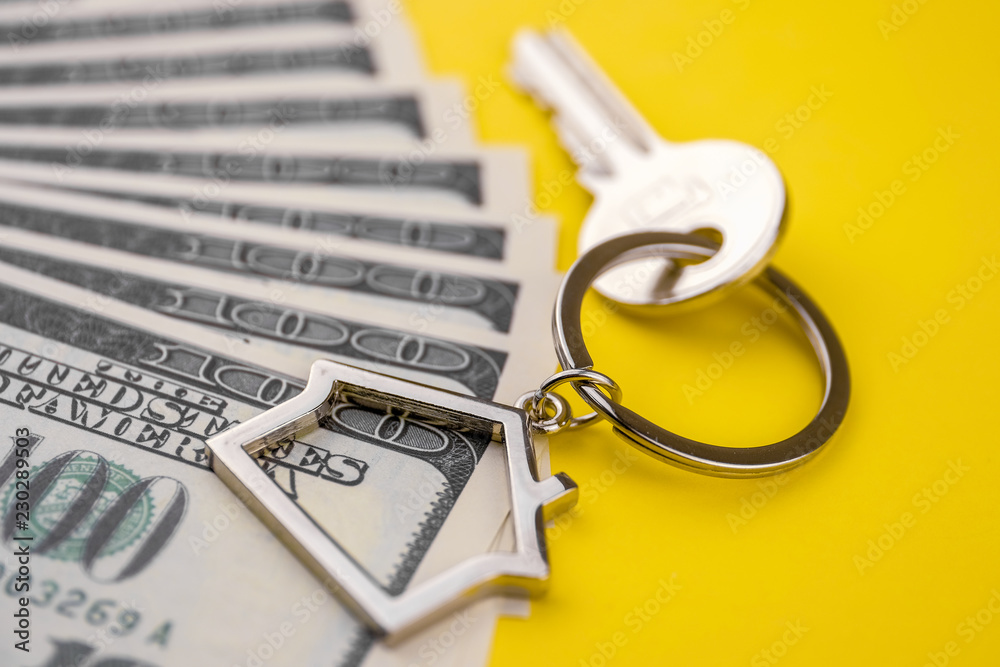 The concept of buying a private house or apartment. Metal key chain in the shape of a house with a metal key lying on the pack of hundred US dollars on a yellow background. Close up. The concept of buying a private house or apartment. Metal key chain in the shape of a house with a metal key lying on the pack of hundred US dollars on a yellow background. Close up.