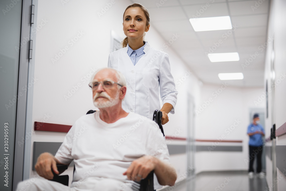Time for a walk. Portrait of serious bearded gentleman sitting in wheelchair while female medic pushing him. Focus on woman Time for a walk. Portrait of serious bearded gentleman sitting in wheelchair while female medic pushing him. Focus on woman