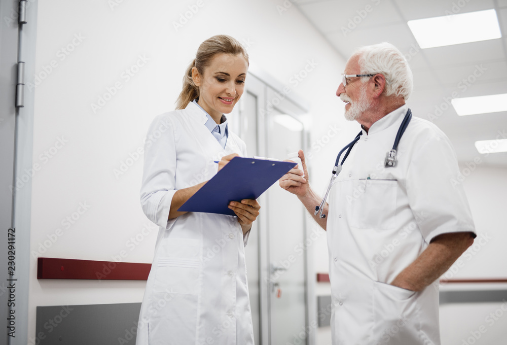 Waist up portrait of old doctor with stethoscope talking with lovely nurse in white lab coat while she writing on clipboard. Bearded gentleman in glasses holding bottle with pills Waist up portrait of old doctor with stethoscope talking with lovely nurse in white lab coat while she writing on clipboard. Bearded gentleman in glasses holding bottle with pills