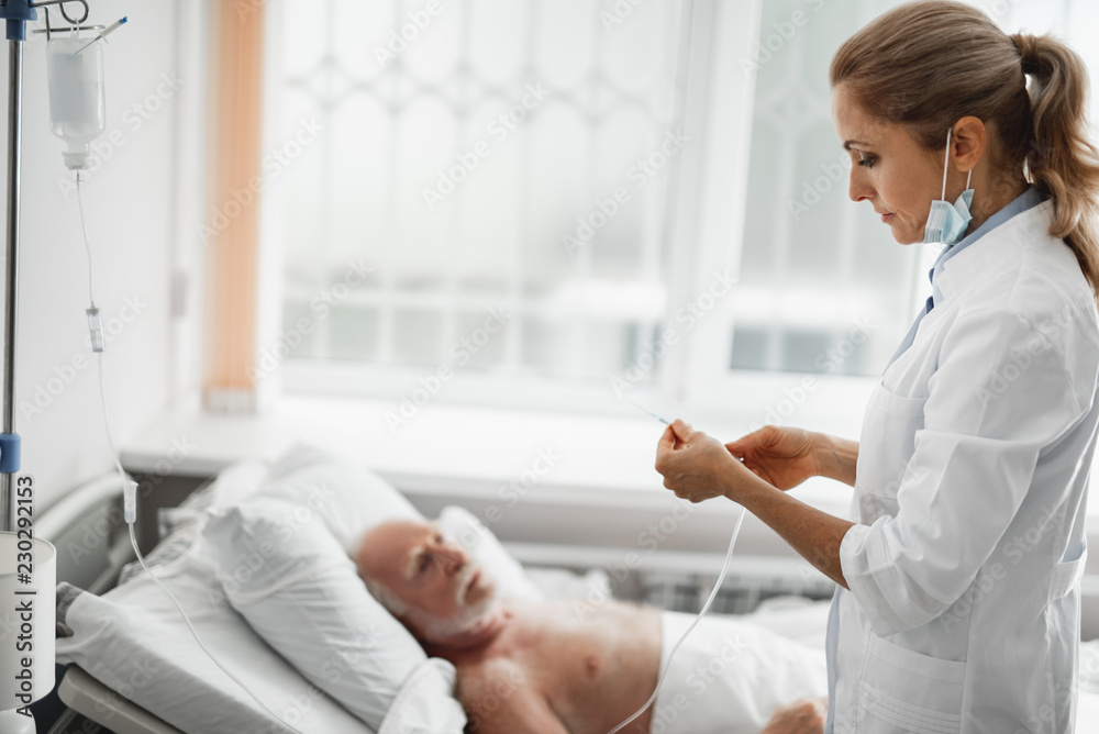 Side view portrait of young lady in white lab coat holding needle for intravenous infusion. Old man lying in bed on blurred background