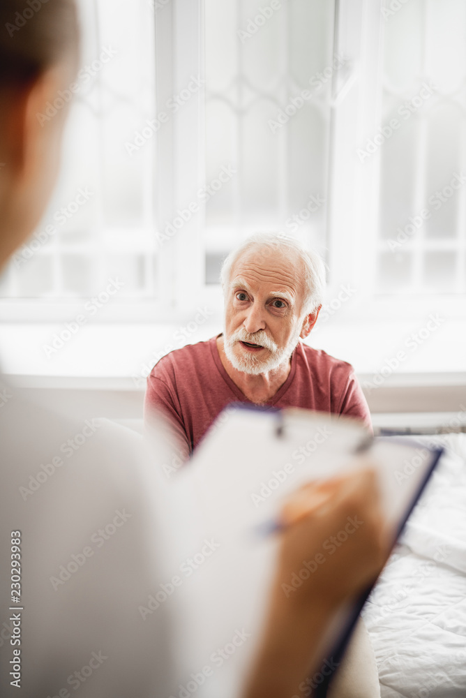 Vertical portrait of bearded gentleman discussing his state of health with female physician while she is listening and noting