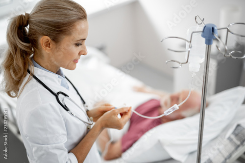 Side view portrait of beautiful young woman in white lab coat checking intravenous drip. Old man resting in hospital room on blurred background photo