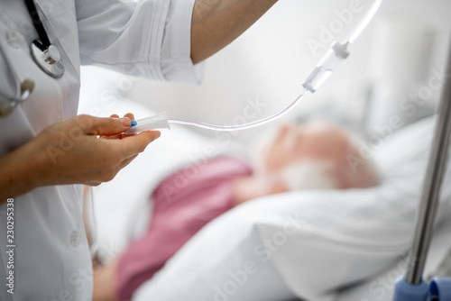 Medical treatment. Close up of female arm regulating IV infusion by using a roller clamp. Old man lying in bed on blurred background photo
