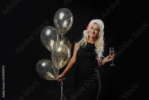 Brightfull expressions portrait of happy emotions  girl in Santa hat celebrating Christmas party on black background. Holds  a glass of champagne and golden balloons. New year holiday concept photo