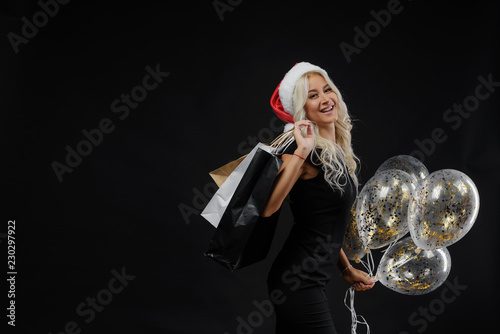 Brightfull expressions portrait of happy emotions  girl in Santa hat celebrating Christmas party on black background. Holds a shopping bags and golden balloons. Sale concept photo