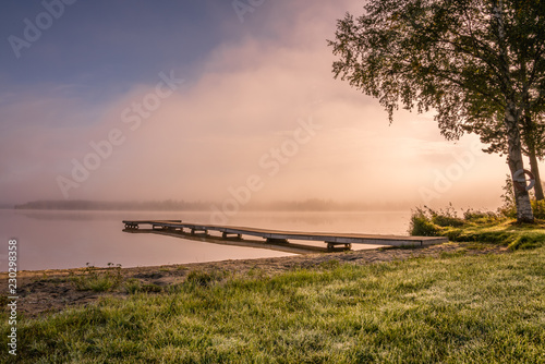 Fototapeta Naklejka Na Ścianę i Meble -  Beautiful golden Sun glows through fog at lake during early sunrise, long wooden bridge, trees, sky reflection in calm water