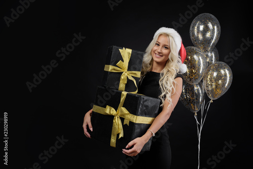 Portrait of beautiful smiling woman in black dress and Santa hat holding stack of gift boxes on the black background. Holding golden balloons and Celebrated a Christmas holiday. Sale concept