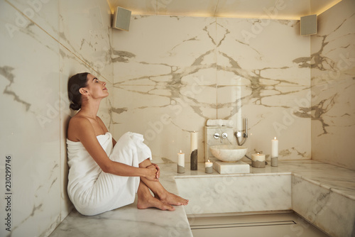 Calm peaceful young woman wearing white towel and sitting next to the wall of comfortable hammam photo