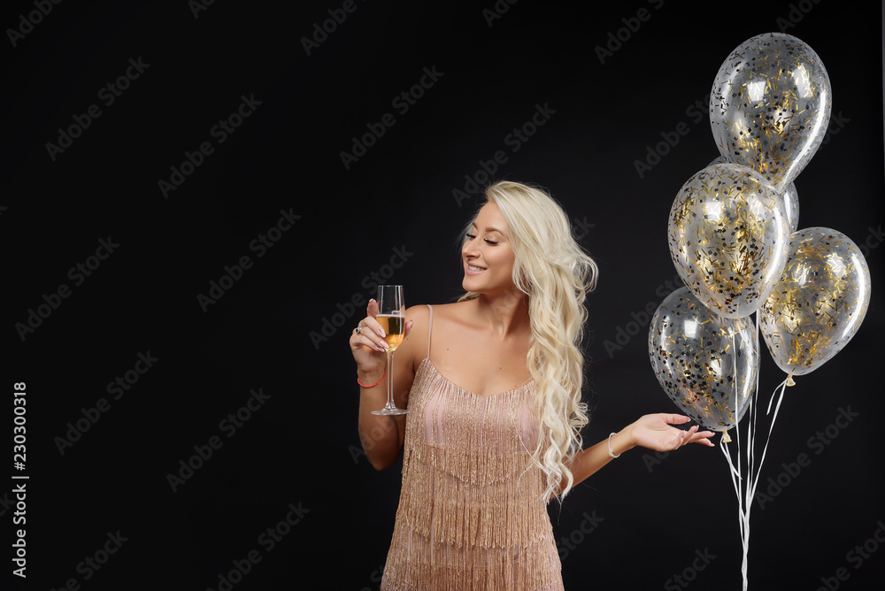 Joyful fashionable young woman in luxury dresses celebrating birthday or Christmas party on black background.Having fun and smiling. Holding present, gift, golden balloons and glass of champagne Joyful fashionable young woman in luxury dresses celebrating birthday or Christmas party on black background.Having fun and smiling. Holding present, gift, golden balloons and glass of champagne