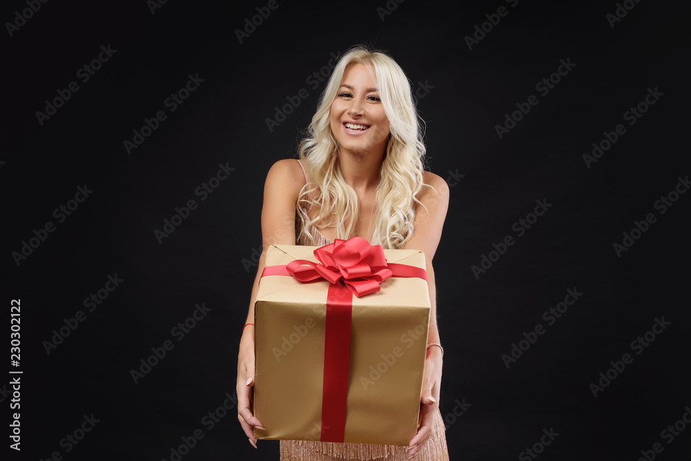 A young blond woman in a festive dress holds in her hands a Christmas gift in a box with a red bow on a black background A young blond woman in a festive dress holds in her hands a Christmas gift in a box with a red bow on a black background