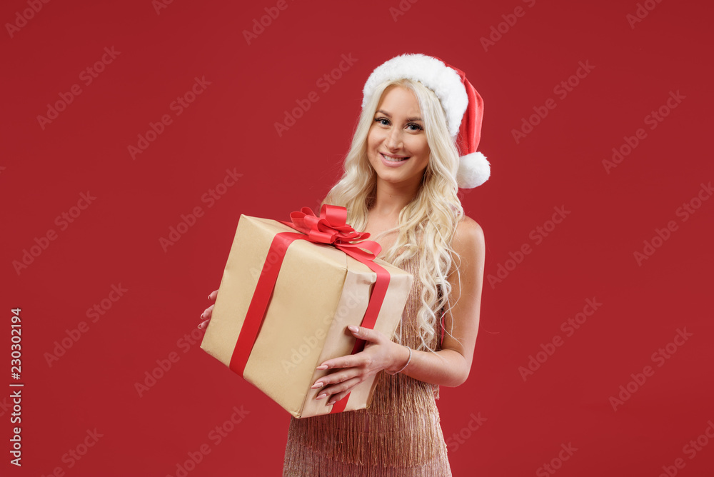 Close up portrait of a happy beautiful smiling woman in gold dress and Santa hat holding a gift box isolated on the red background. Christmas celebrate and surprise Close up portrait of a happy beautiful smiling woman in gold dress and Santa hat holding a gift box isolated on the red background. Christmas celebrate and surprise
