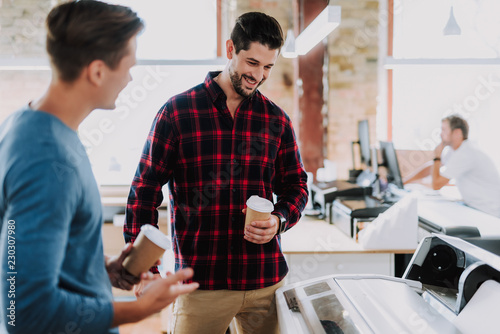 Cheerful man drinking coffee while standing with his male colleague near a printer