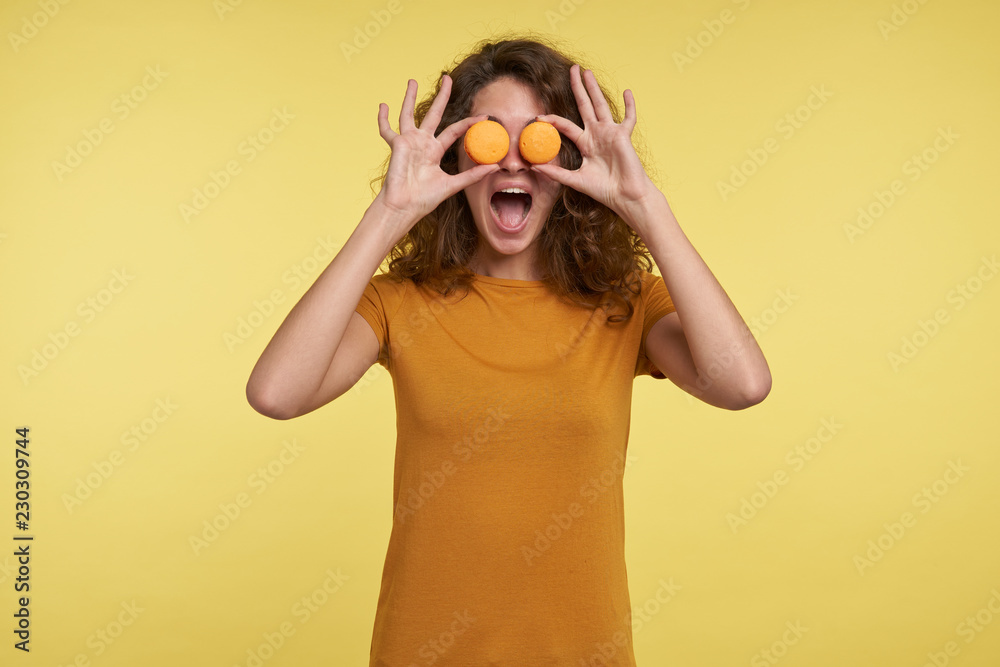 Funny smiling young brunette woman shows macaroons in front of the eyes, isolated over yellow background