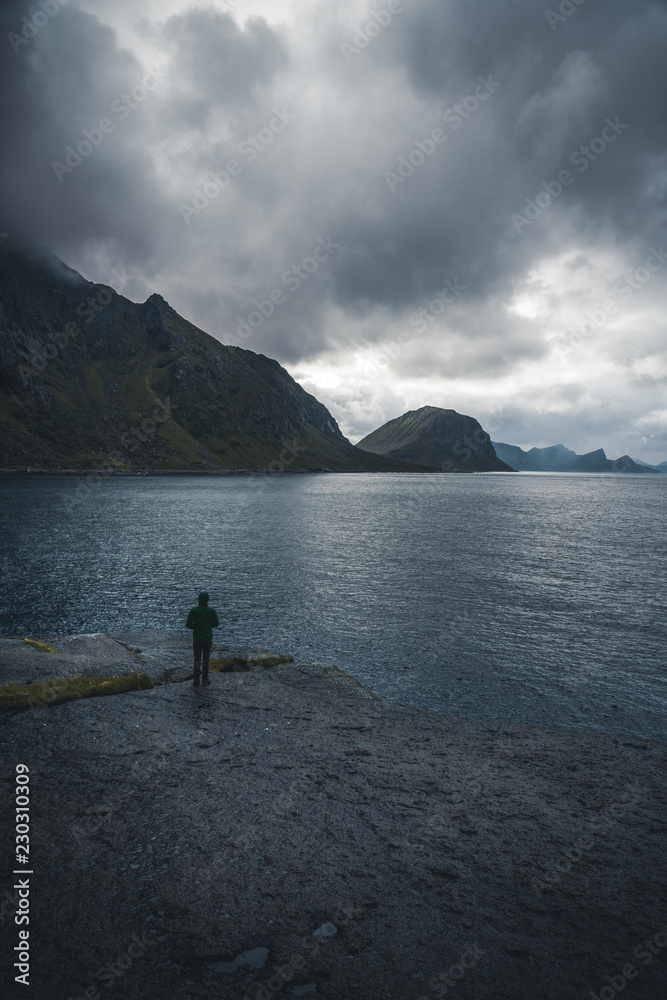 Lonely man standing in front of the atlantic ocean and looking at the stormy sea. Silhouette of seascape with alone man on the rocks at sunset sky. Photo taken in Lofoten Islands, Norway.